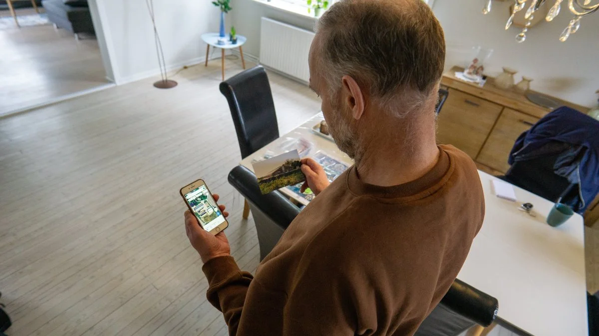 Johannes Linschoten står her med sin telefon med gårdens beliggenhed og et fysisk billede af den gamle gård, som stadig volder ham mange frustrationer. Foto: Alexander Dornwirth.