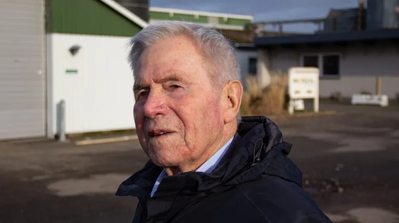 Aage Jakobsen står der, hvor han i 1957 etablerede sig på 8 hektar sandjord. Ejendommen udviklede sig hurtigt til en af landets største svinebesætninger. I baggrunden ses hans første modulbyggede stald fra 1966, som var startskuddet til hans mangeårige karriere som byggematador af stalde, fabriksbygninger og sportshaller. Foto: Anne Wolfenberg